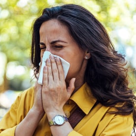 Portrait of woman outside near trees with napkin blowing nose.