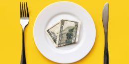 A hundred-dollar bill on a white plate between a fork and a knife on a yellow tablecloth.